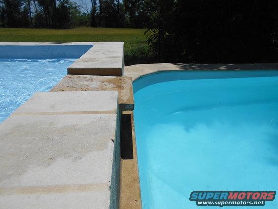 Two swimming pools with concrete decking, one with blue water, the other with clear water. Trees and grass in background. Supermotors.net logo in corner