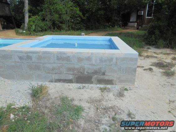 Concrete block spa with blue water, surrounded by dirt and grass. A wooden structure is visible in the background