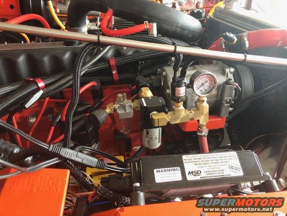 Engine bay with orange components, black hoses, and a pressure gauge. A warning label is visible