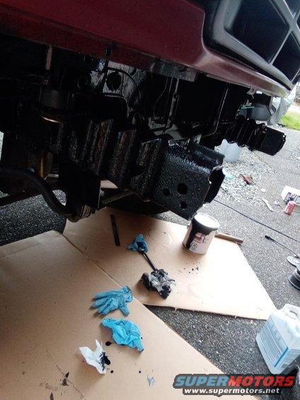 Frame and bumper area of a vehicle being painted black. Gloves and paint can are on cardboard