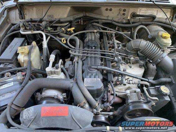 2000 Jeep Cherokee engine bay with visible hoses, belts, and engine components. Text on a red label is partially visible