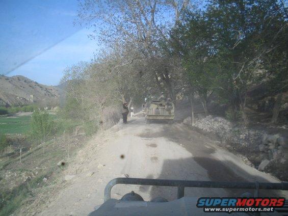 Military truck drives on a dusty dirt road through a rural landscape with trees and hills