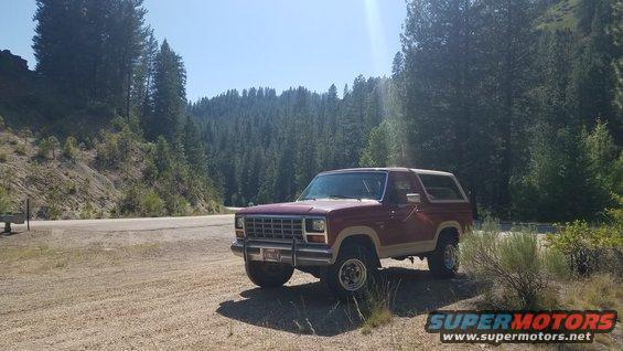 1985 Ford Bronco parked on dirt road, trees and hills behind