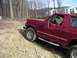 1994 Ford Bronco - A maroon Ford Bronco is seen driving on a dirt path with trees and buildings in the background.