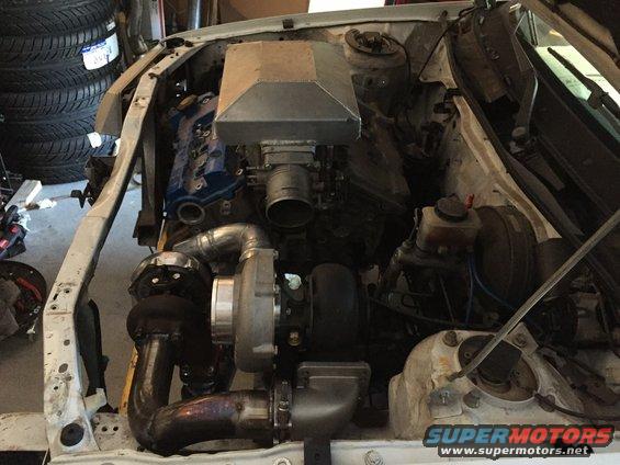Engine bay with turbocharger and custom intake, white car body, tires stacked in background