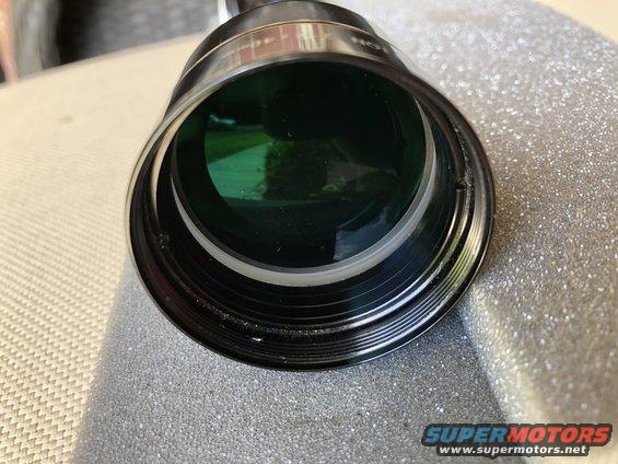 Close-up of a camera lens with green tint and metallic threading, resting on a textured surface