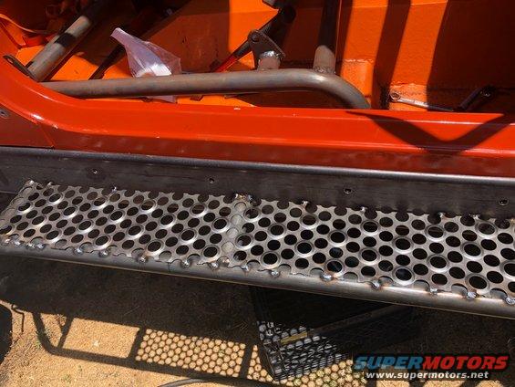 Close-up of custom metal rock sliders with perforated steps on an orange vehicle's side, showing interior roll cage bars