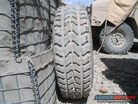 Close-up of a large tire next to a sandbag barrier, with a military vehicle visible in the background