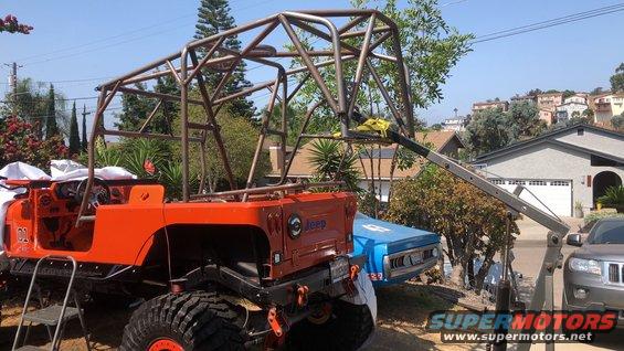Orange Jeep with a large metal roll cage being worked on, with a blue car and a gray SUV nearby