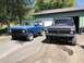 1978 Ford Bronco - Two Ford Broncos, a blue first-generation model and a matte black one, are parked outside a garage.