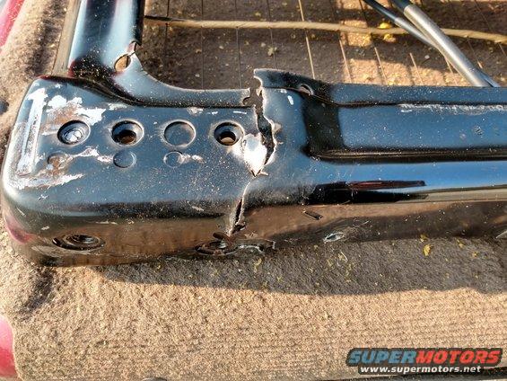 Close-up of a damaged black metal seat frame with holes and a large crack. It's resting on a tan fabric surface