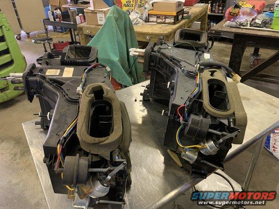 Two car heater core assemblies are on a metal table in a workshop. One is in the foreground, the other is behind it