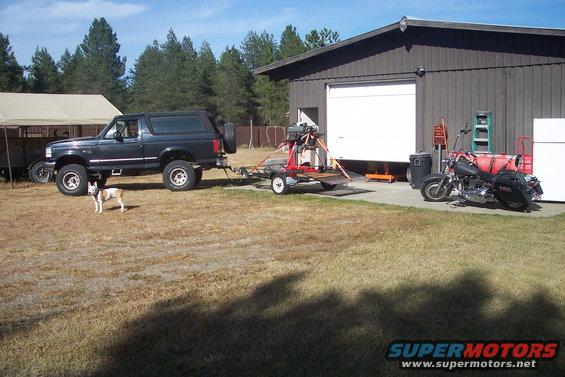 Black Ford Bronco towing a trailer with equipment, parked next to a garage with a motorcycle