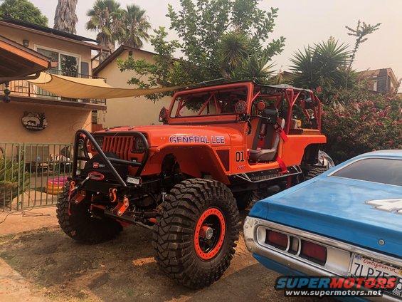 Orange Jeep Wrangler with 'GENERAL LEE' and '01' graphics, roll cage, large off-road tires, and winch. A blue car is parked next to it