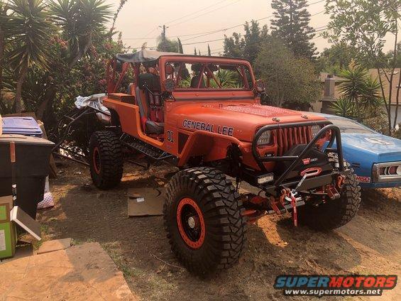 Orange Jeep Wrangler with "GENERAL LEE" and "01" on the hood, large off-road tires, and a winch on the front bumper. A blue car is parked next to it