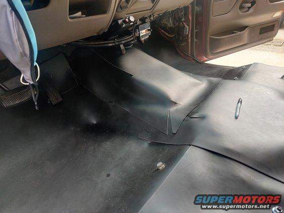 Interior floor of a vehicle with black rubber matting. Shows pedals, dashboard base, and door panel