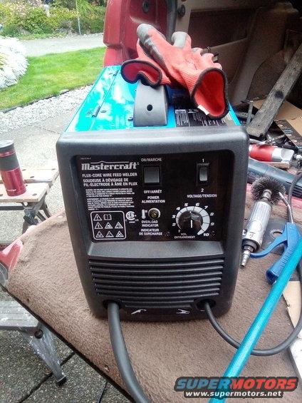 Mastercraft wire feed welder with orange gloves on top, sitting on a brown blanket inside a vehicle