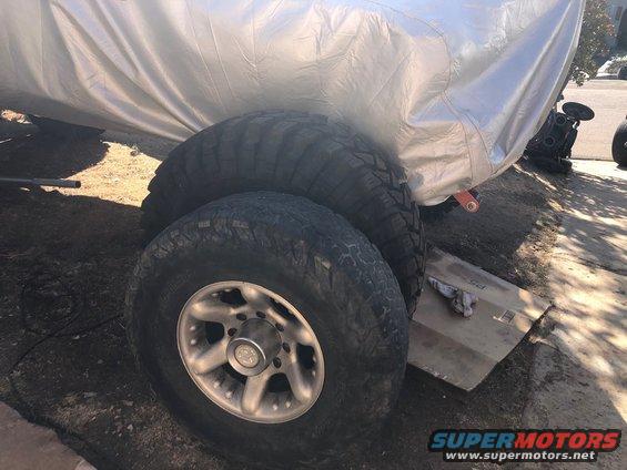 Two large off-road tires stacked, bottom tire on a silver rim, under a silver tarp.