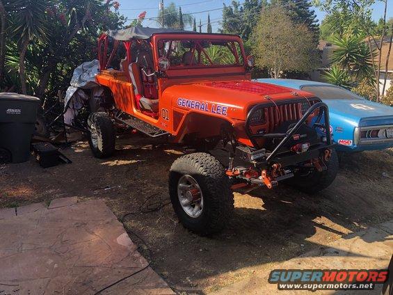 Orange Jeep Wrangler "General Lee" with "01" on the side, parked outdoors next to a blue car.