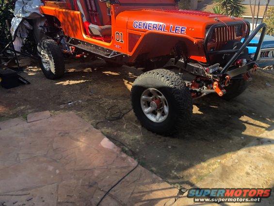 Orange Jeep Wrangler with "General Lee" text, off-road tires, and black roll cage parked outdoors.