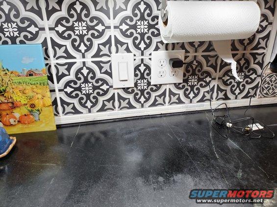Kitchen counter with patterned tile backsplash, paper towel holder, light switch, and electrical outlets. A fall-themed picture is on the left
