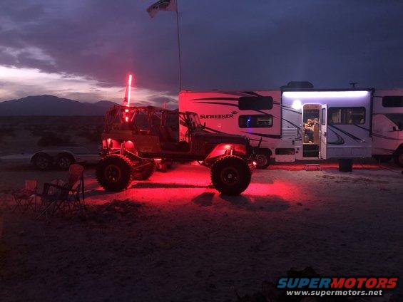 Modified Jeep Wrangler with red underglow and off-road tires, parked next to a recreational vehicle at dusk
