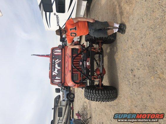 Orange Jeep Wrangler with a man in an orange shirt and hat standing next to it. The Jeep has a roll cage and large tires