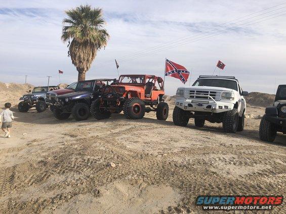 Several modified off-road vehicles parked on sandy terrain, with a palm tree and flags in the background