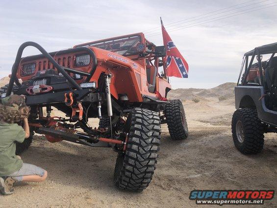 Orange 1992 Jeep Wrangler with custom bumper, winch, and large off-road tires. Another Jeep is parked to the right. A Confederate flag flies from the Wrangler