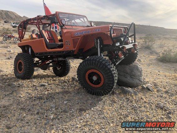 Orange Jeep Wrangler with roll cage, large tires, and "GENERAL LEE" text on fender, parked on rocky terrain