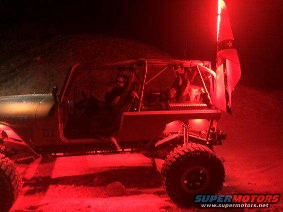 Modified Jeep with roll cage and red underglow, flag on back, at night