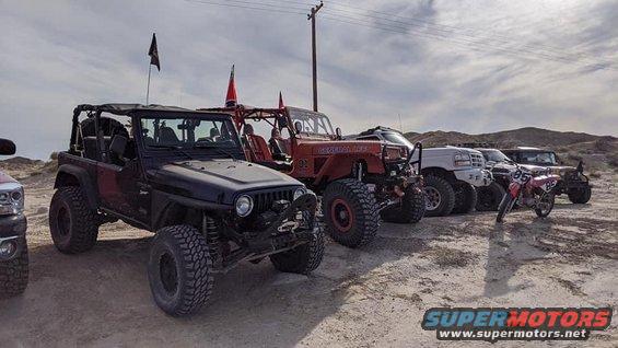 Black Jeep Wrangler with large tires parked on dirt, other off-road vehicles and a dirt bike behind it
