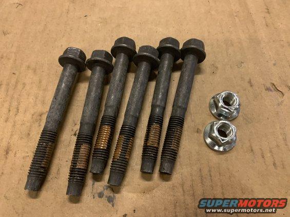 Six used bolts and two nuts on a brown surface. The bolts have hexagonal heads and threaded ends with some rust