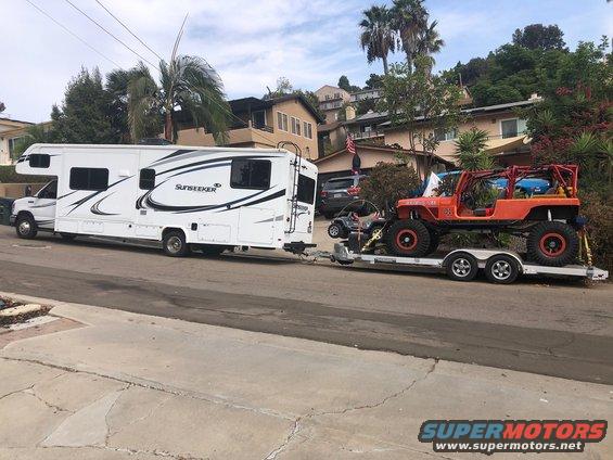 Orange Jeep Wrangler on a dual-axle trailer, towed by a white RV. A small ATV is also on the trailer