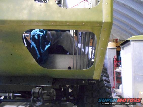 Side view of a green vehicle body with a large opening showing interior components and some blue fabric. The vehicle appears to be in a workshop