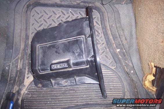 Black plastic heater vent cover on a ribbed floor mat. Text 'VENT' is visible