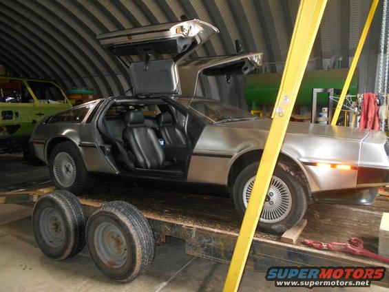 Silver DeLorean DMC-12 with gullwing doors open, on a trailer, with a yellow support beam. Another vehicle is visible behind it