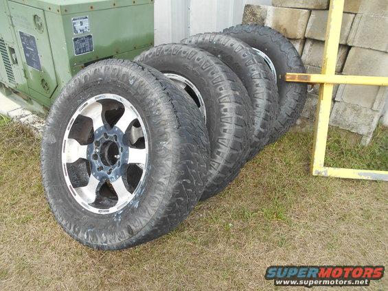Four off-road tires with silver and black rims on grass, next to a green generator and concrete blocks
