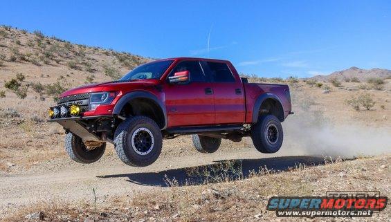 Red pickup truck mid-air jump over a dirt road in a desert landscape. It has an aftermarket bumper with lights