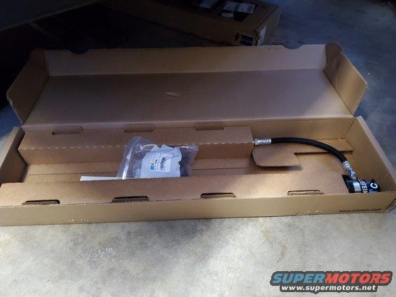 Cardboard box containing a black hose with metal fittings and a bag of hardware, with text 'BILSTEIN' visible