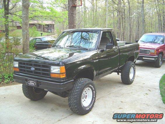 Lifted black 1989 Ford Ranger pickup truck with off-road tires parked on a driveway. Red SUV in background