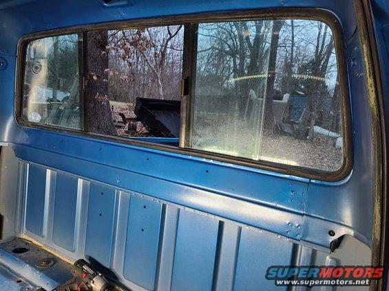 Inside of a blue truck cab, looking out the rear window at trees and a black truck bed