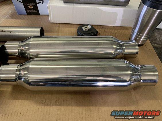 Two shiny exhaust mufflers sit on a cardboard surface next to a metal cup and some boxes