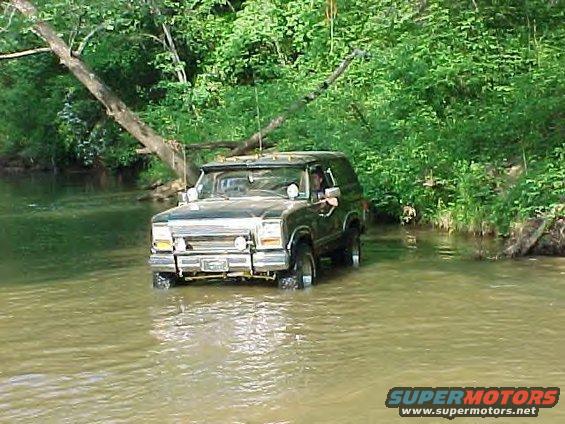 Dark green 1987 Ford Bronco driving through shallow water, front view