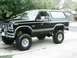 1984 Ford Bronco - This lifted black Ford Bronco is parked on a concrete driveway with trees and houses visible in the background.