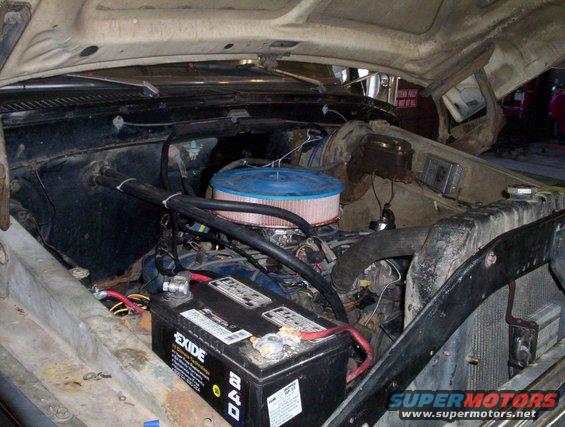 Engine bay of a dark-colored vehicle with an open hood, showing a battery, air filter, and hoses