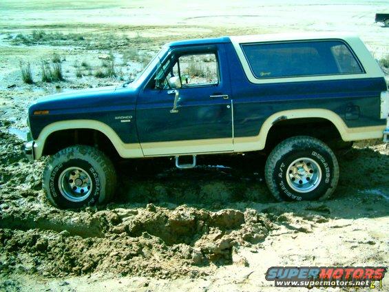 Blue and tan Ford Bronco stuck in mud, side view. Appears to be from the early 1980s