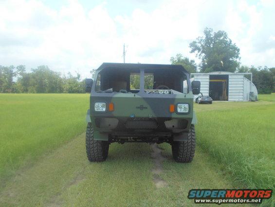 Front view of a camouflaged military truck with its hood open. A sports car is parked near a metal shed in the background