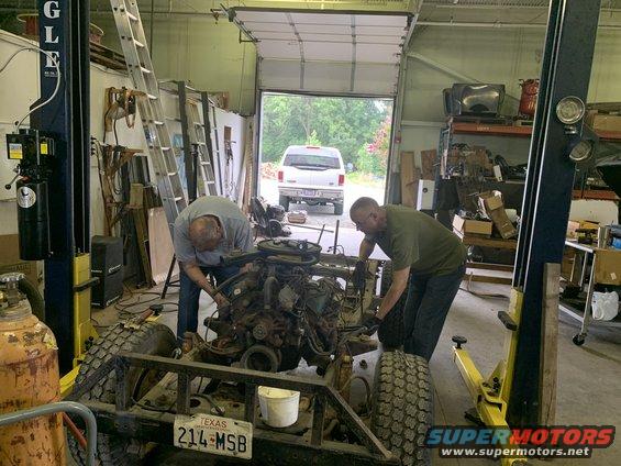 Two men working on a vehicle frame and engine in a garage; Texas license plate 214-MSB visible