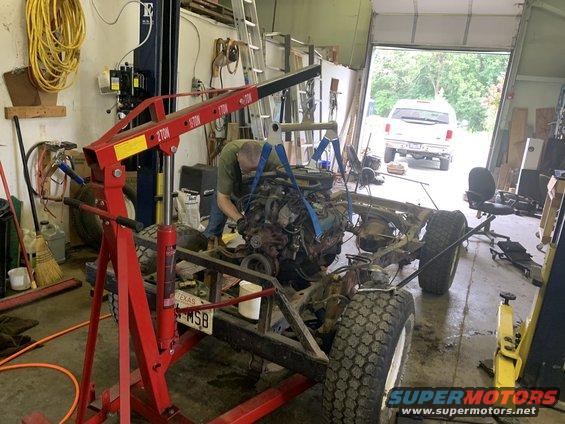 Jeep CJ frame on the ground, engine being lifted out by an engine hoist. Another vehicle is parked outside the garage door